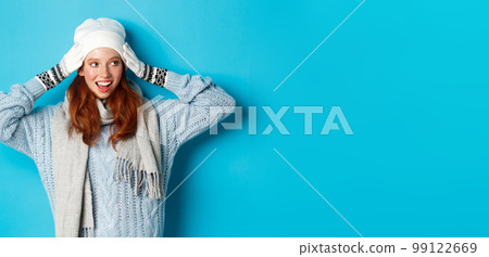 Winter and holidays concept. Happy redhead girl in beanie hat, scarf and gloves looking right and smiling, standing against blue background 99122669