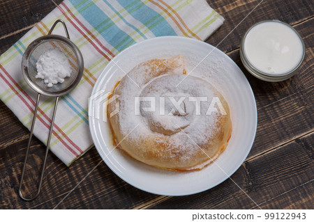 On a wooden table a vertuta sprinkled with powdered sugar and a towel, next to it is a bowl with white sauce On a wooden table a vertuta sprinkled with powdered sugar and a towel, next to it is a bowl with white sauce 99122943
