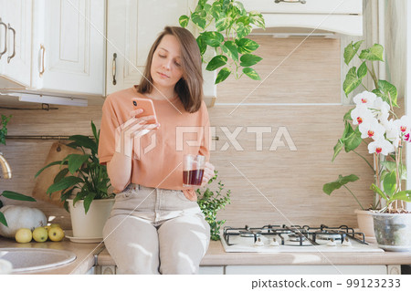 woman is sitting on table in kitchen chatting in a smartphone and drinking coffee brewing pourover woman is sitting on table in kitchen chatting in a smartphone and drinking coffee brewing pourover 99123233
