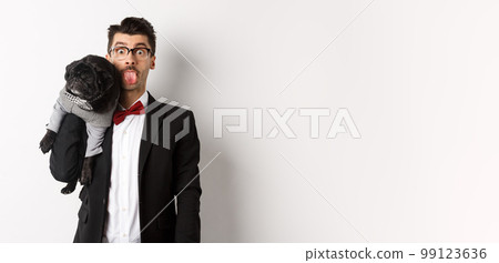 Funny young man in party suit, showing tongue and holding cute black pug on shoulder, celebrating with pet, standing over white background Funny young man in party suit, showing tongue and holding cute black pug on shoulder, celebrating with pet, standing over white background 99123636