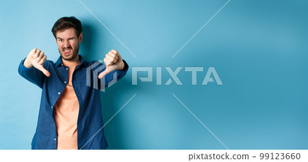 Disgusted guy express negative opinion, showing thumbs down and tongue, frowning upset, disapprove something bad, standing on blue background Disgusted guy express negative opinion, showing thumbs down and tongue, frowning upset, disapprove something bad, standing on blue background 99123660