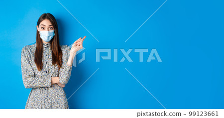 Covid-19, social distancing and healthcare concept. Excited caucasian woman in medical mask raise eyebrows, pointing right at interesting promotion, blue background Covid-19, social distancing and healthcare concept. Excited caucasian woman in medical mask raise eyebrows, pointing right at interesting promotion, blue background 99123661