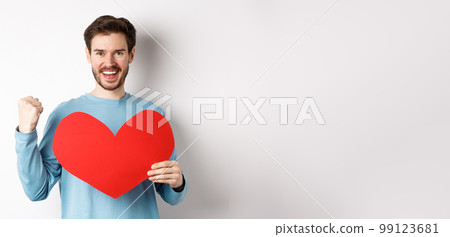 Cheerful man determined to find love on Valentines day, making fist pump success gesture and holding big red romantic heart, standing over white background 99123681