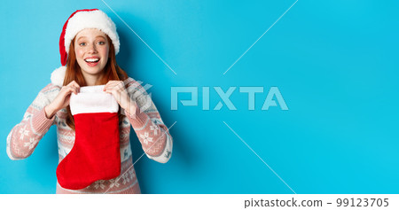 Beautiful redhead girl in santa hat open Christmas stocking and looking surprised, receiving xmas gift, standing over blue background 99123705