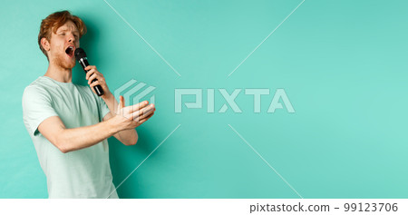 Passionate redhead man in t-shirt singing serenade with microphone, looking aside at karaoke and gesturing, standing over mint background 99123706
