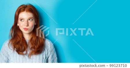 Close-up of cute redhead girl thinking and looking left, have an idea, standing against blue background Close-up of cute redhead girl thinking and looking left, have an idea, standing against blue background 99123710