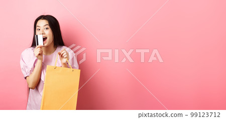 Shopping. Excited beautiful asian woman buying in stores, holding paper bag and kissing plastic credit card, standing against pink background Shopping. Excited beautiful asian woman buying in stores, holding paper bag and kissing plastic credit card, standing against pink background 99123712