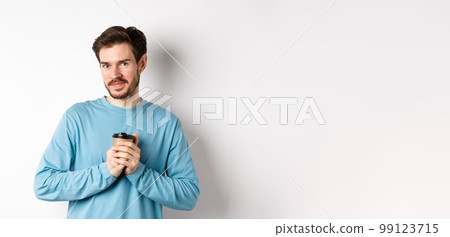 Handsome caucasian man warm-up hands with cup of coffee, smiling at camera and looking tenderly, standing on white background 99123715