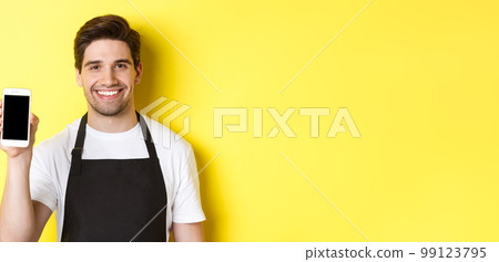 Close-up of handsome waiter in black apron showing smartphone screen, recommending app, standing over yellow background 99123795