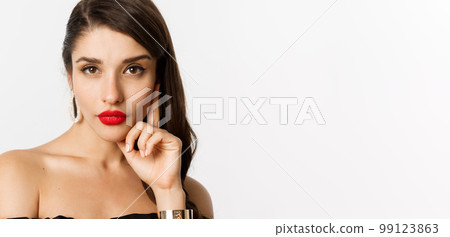 Fashion and beauty concept. Close-up of elegant beautiful woman in black dress, evening makeup and red lipstick, looking sassy at camera, standing over white background Fashion and beauty concept. Close-up of elegant beautiful woman in black dress, evening makeup and red lipstick, looking sassy at camera, standing over white background 99123863