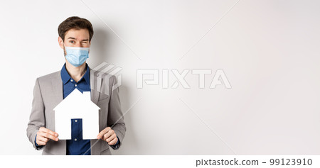 Real estate and covid-19 concept. Cheerful young man in medical mask and suit winking at camera, advertising new flats, showing paper house cutout 99123910