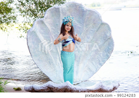 A cute girl in a mermaid costume stands outdoor in a large sea shell, holds her thumbs up 99124283