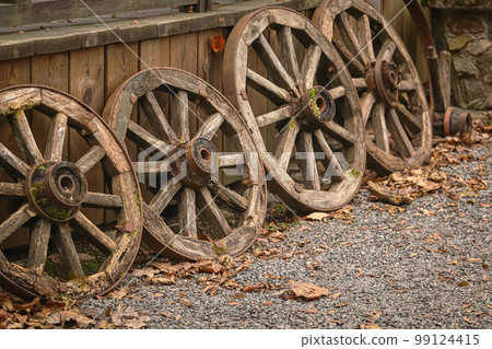 Old wooden wheels arranged in a row near wooden plank elevation on pebble road with autumn leaves Old wooden wheels arranged in a row near wooden plank elevation on pebble road with autumn leaves 99124415