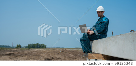 Caucasian male builder in hardhat sits on floor slabs and uses laptop at construction site. Caucasian male builder in hardhat sits on floor slabs and uses laptop at construction site. 99125348