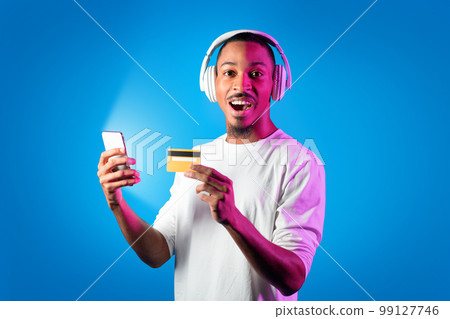 Excited young black guy using smartphone, headphones and credit card 99127746