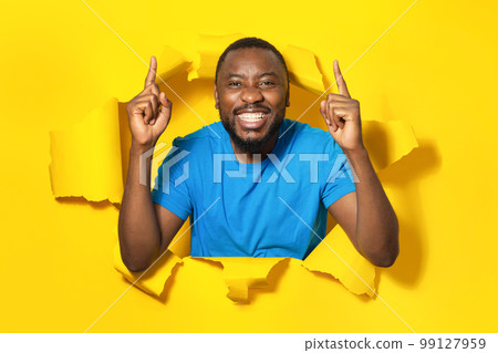 Portrait of excited african american man pointing fingers up and smiling, standing in torn paper hole yellow background Portrait of excited african american man pointing fingers up and smiling, standing in torn paper hole yellow background 99127959