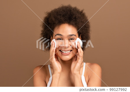 Glad young african american curly lady with perfect skin in white top puts cotton pads to face, has fun Glad young african american curly lady with perfect skin in white top puts cotton pads to face, has fun 99127992