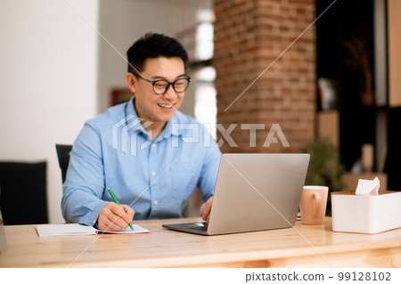 Happy asian man looking at laptop screen and writing in notebook, sitting at workplace at home office, selective focus 99128102