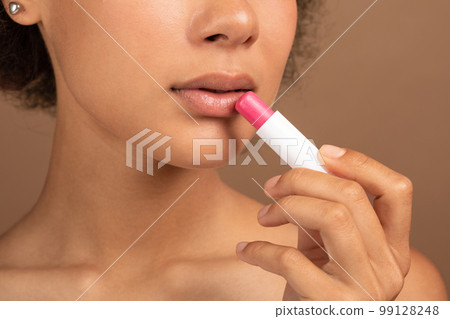 Serious young black curly lady with perfect skin applies lipstick on lips for moisturizing 99128248