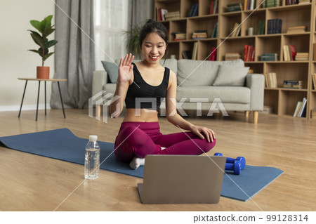 Online domestic training. Happy young asian lady waving to webcam, sitting on yoga mat in front of laptop, free space 99128314