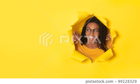 Excited african american lady in glasses posing in torn paper, looking at free space through hole on yellow background 99128329