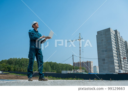 A male builder in a helmet stands against the background of a construction site with a laptop in his hands.  99129663