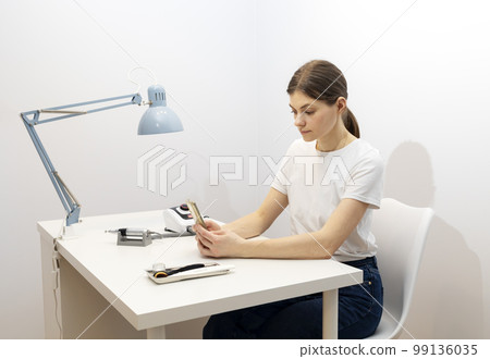 Nail beautician hold gadget, looking at smartphone, sitting at workstation in nail beauty treatment salon. Professional tools are on white table. Studying manicurist. Closeup, horizontal 99136035