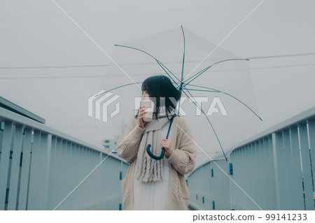 Photo of a woman holding an umbrella on an overpass and drinking coffee while waiting for a meeting 99141233