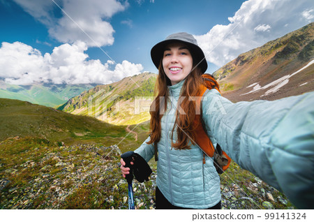 Smiling young woman takes a selfie on a mountain peak, with a backpack on a sunny day. Tourist hiking in the mountains Smiling young woman takes a selfie on a mountain peak, with a backpack on a sunny day. Tourist hiking in the mountains 99141324