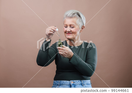 Portrait of smiling senior woman holding skincare serum drops in studio.Concept of beauty and skincare. 99144661