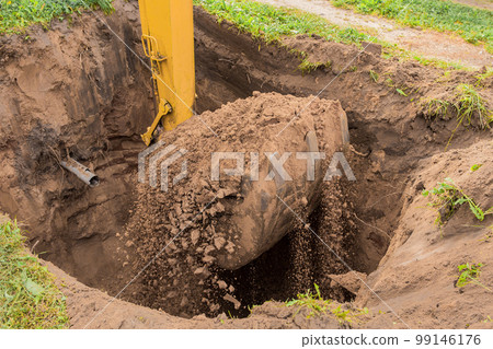 A bucket of excavator with a pile of earth digs a hole on the construction site. Industrial work 99146176