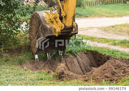 A bucket of a bulldozer digs the ground with a grass close-up in an industrial area. Excavation work on the construction site 99146179