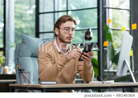 Serious young man programmer, freelancer sitting at the desk in the office, working at the computer. Holds a phone in his hands, uses a gadget. Writes messages, checks mail, chats. 99146369