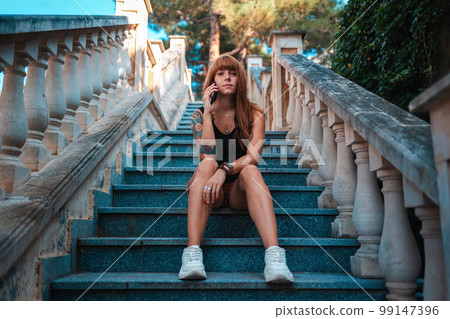 A young woman with tattoos is sitting on the stairs and talking on the phone. Bottom view. Communication concept A young woman with tattoos is sitting on the stairs and talking on the phone. Bottom view. Communication concept 99147396