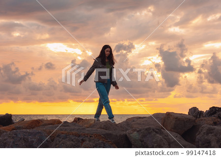 A young woman walks alone on the sea rocks. The dramatic sunset in the background. The concept of psychology and mental health 99147418