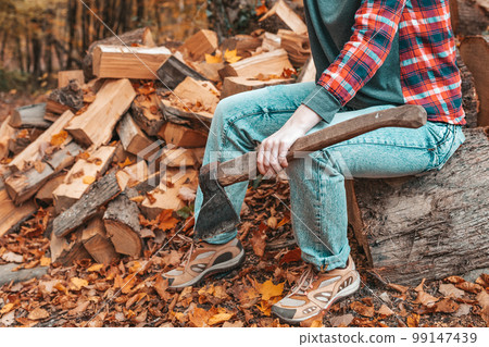 Preparation for the heating season. A young woman with an axe in her hands sits on a log. In the background, a pile of firewood. Close up 99147439