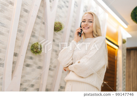 Low-angle view of beautiful young woman talking on smartphone standing in hotel lounge, looking at camera. Cute blonde female using phone in lobby. Pretty lady having call phone standing in doorway Low-angle view of beautiful young woman talking on smartphone standing in hotel lounge, looking at camera. Cute blonde female using phone in lobby. Pretty lady having call phone standing in doorway 99147746