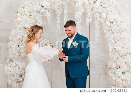 happy bride and groom exchange rings in an arch of artificial flowers. 99148811