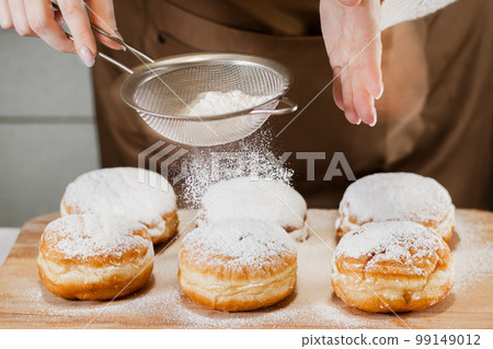 Woman chef prepares fresh donuts in her bakery. Cooking traditional Jewish Hanukkah Sufganiyot. Small business concept. Hands sprinkle Berliners with powdered sugar. Woman chef prepares fresh donuts in her bakery. Cooking traditional Jewish Hanukkah Sufganiyot. Small business concept. Hands sprinkle Berliners with powdered sugar. 99149012