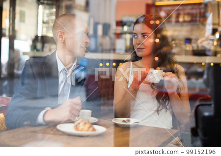 An elegant couple are talking and drinking coffee at table in a cafe. View from street An elegant couple are talking and drinking coffee at table in a cafe. View from street 99152196