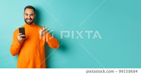 Photo of satisfied young man in orange sweater, showing thumb-up after reading on mobile phone, standing pleased against turquoise background 99158684