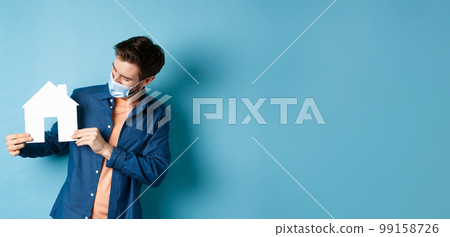 Real estate and quarantine concept. Young caucasian man in medical mask looking at paper house cutout, standing on blue background Real estate and quarantine concept. Young caucasian man in medical mask looking at paper house cutout, standing on blue background 99158726