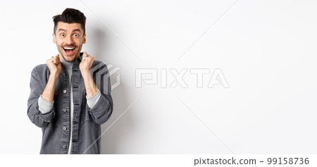 Excited young man jumping from happiness, smiling amazed and holding hands pressed to chest, standing on white background 99158736