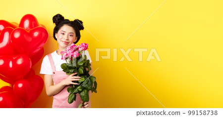 Happy Valentines day. Beautiful asian woman dressed for romantic date, holding bouquet of flowers and smiling. Girl with roses standing near heart balloons, yellow background Happy Valentines day. Beautiful asian woman dressed for romantic date, holding bouquet of flowers and smiling. Girl with roses standing near heart balloons, yellow background 99158738