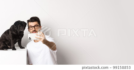 Image of handsome young man taking selfie with cute black dog on smartphone, posing with pug over white background 99158853