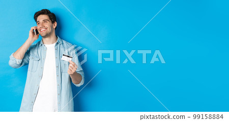 Smiling man call bank support, talking on mobile phone and holding credit card, standing against blue background 99158884