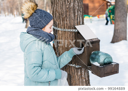 The girl wants to put the seeds in the bird feeder on the tree in the winter forest. The girl wants to put the seeds in the bird feeder on the tree in the winter forest. 99164889