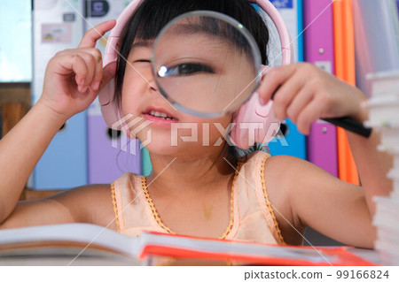 Cheerful little girl in headphones reading a book with a magnifying glass sitting at the table in her room at home. 99166824