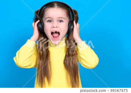 Portrait of a girl on a blue background in a yellow sweater, a little girl with wireless headphones. 99167875