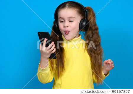 Portrait of a little girl with a phone and wireless headphones, a child in a yellow sweater on a blue background. 99167897